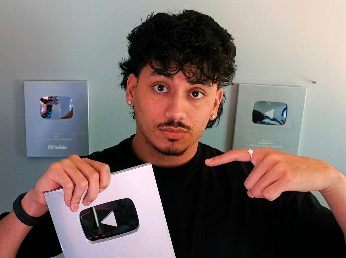 Brem holding up a YouTube Silver Creator Award and pointing at it, with two more plaques on the wall behind him